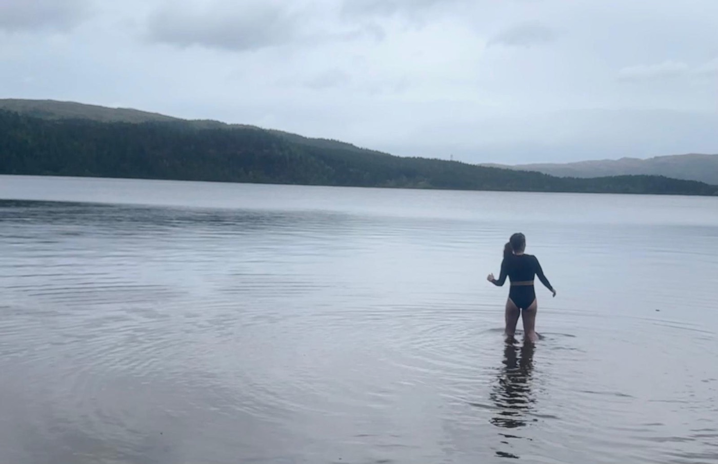 Aimee wading into a still lake below wooded hills.